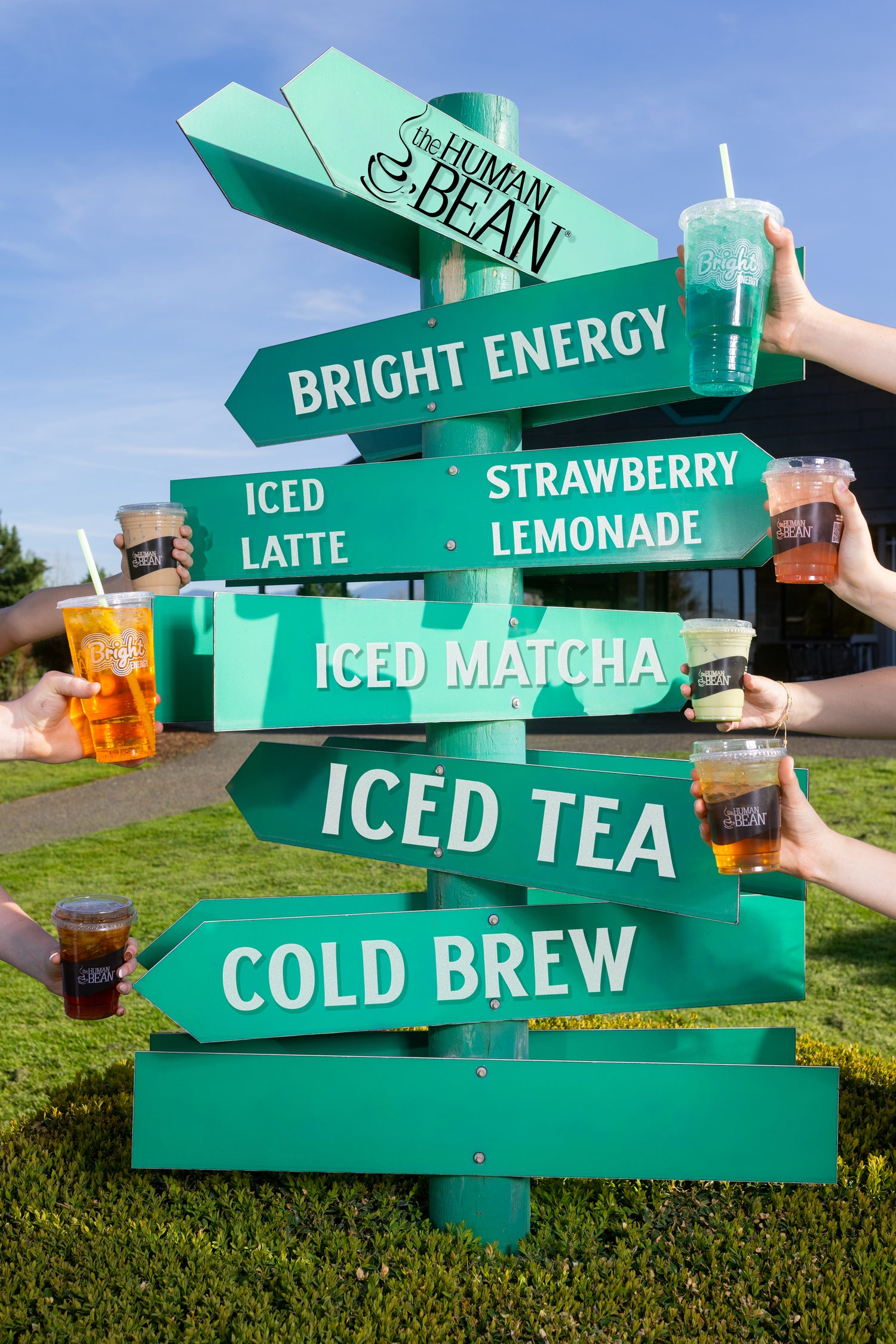 Green signpost with coffee drink options like 'Bright Energy', 'Strawberry Lemonade', and 'ICED TEA' with people holding drinks around it.